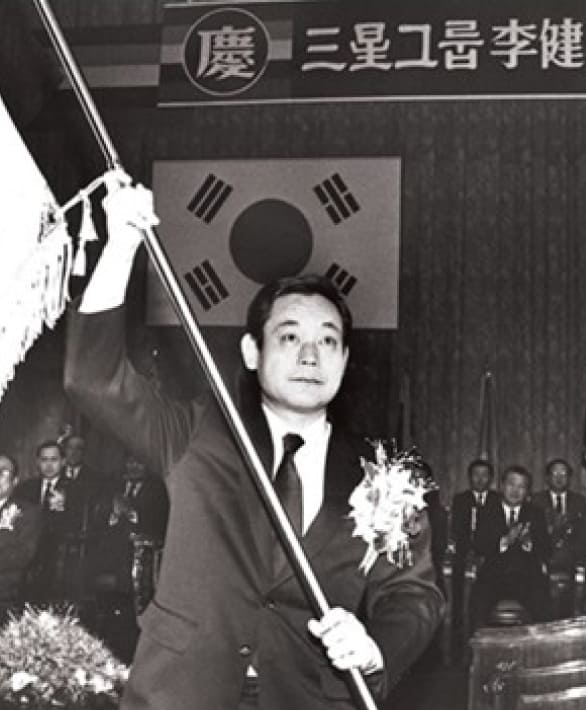 A black-and-white photo of Chairman Lee Kun-hee waving a Samsung flag at his inauguration as Chairman in December 1987.